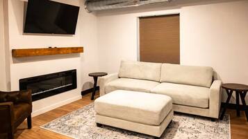 A living room with a sofa, ottoman, TV, fireplace, and a round table.