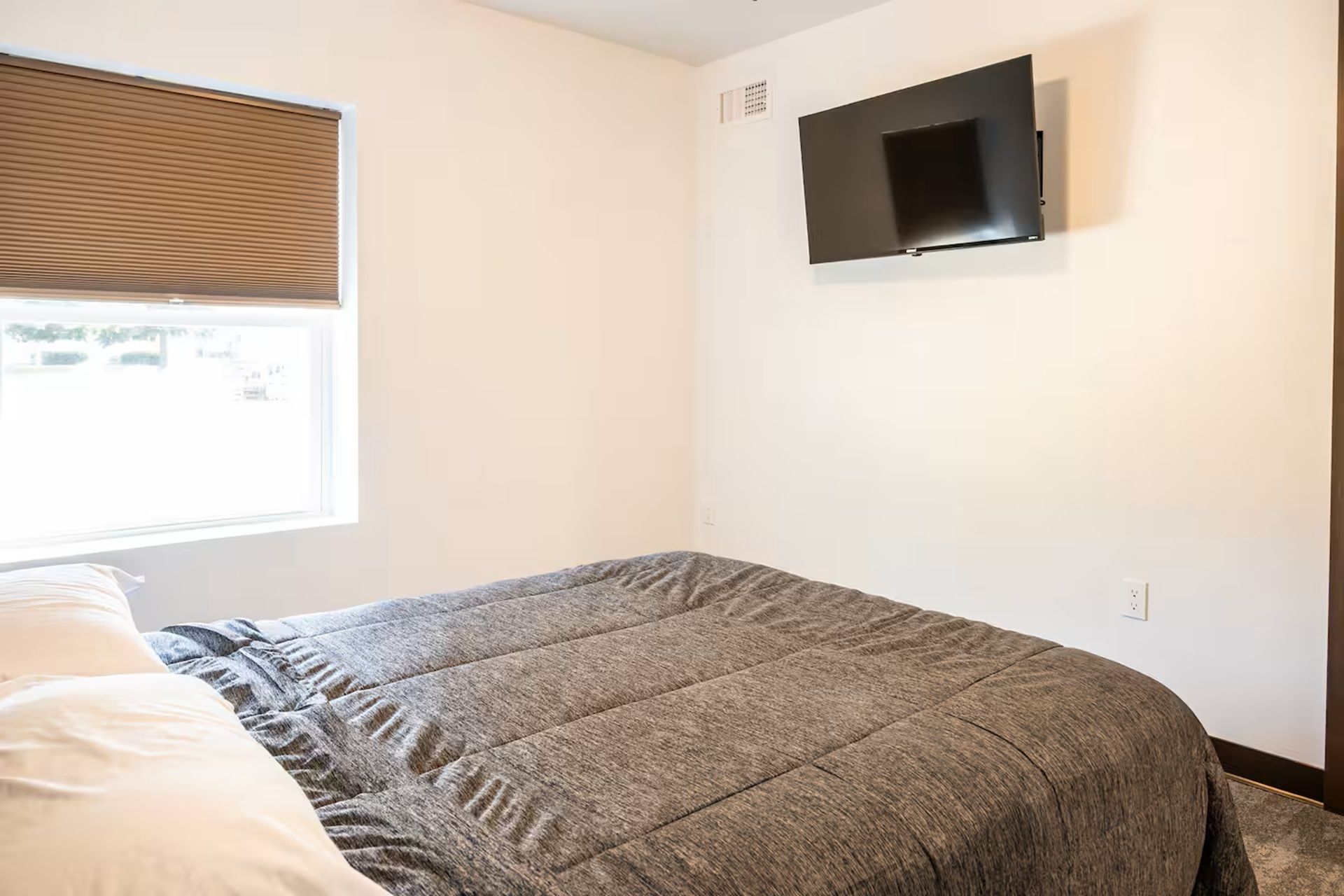 Un dormitorio con cama, ventana con persianas, televisor de pared y toma de corriente.