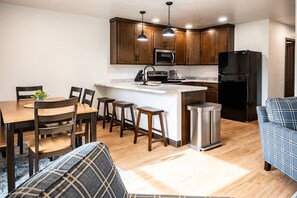 A modern kitchen with wooden cabinets, a central island, a black refrigerator, and a dining area with a table and chairs. - Exclusive Full-Complex Retreat: Reserve All 7 Suites & Common Areas (Brillion)