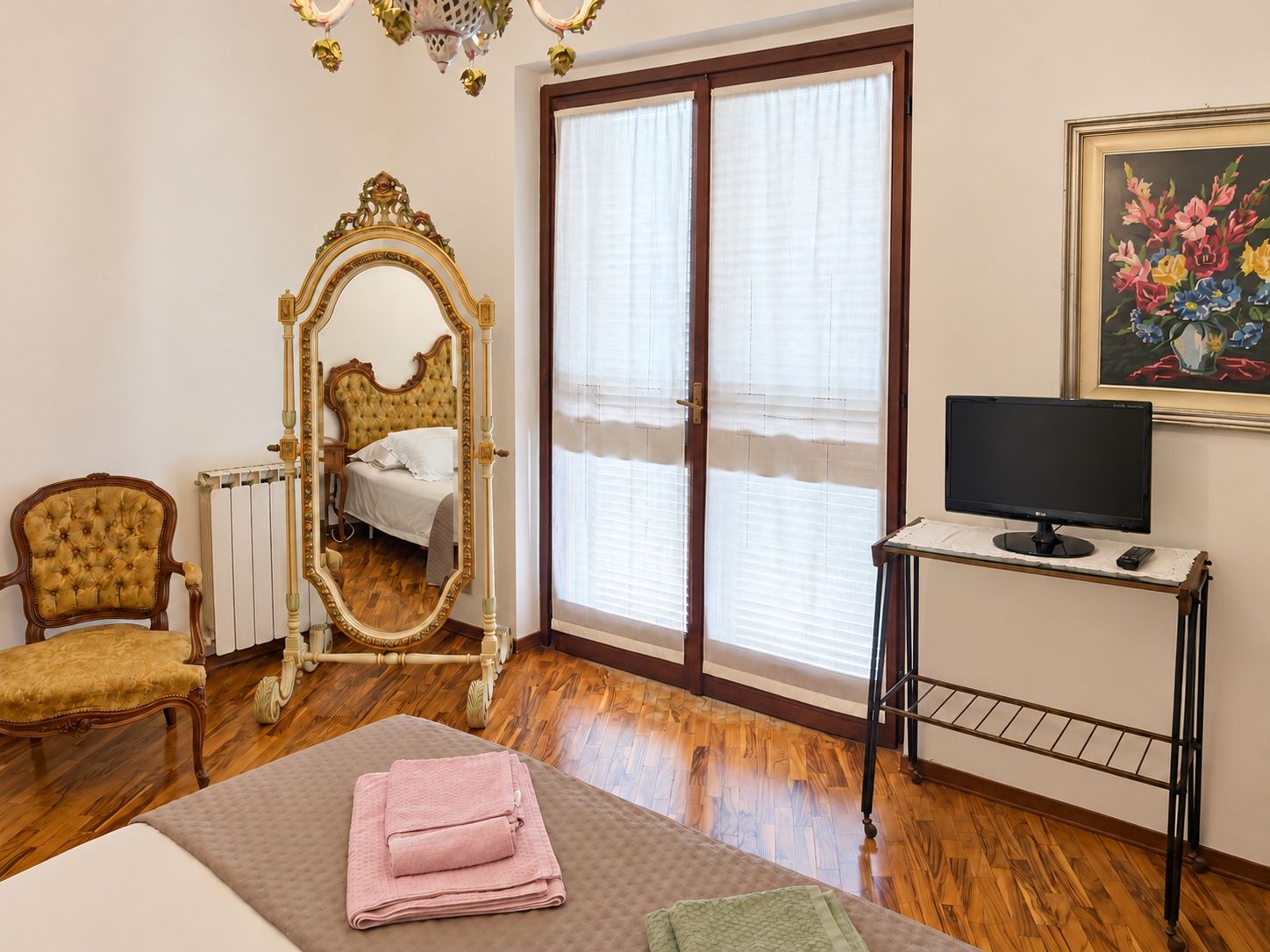 Une chambre à coucher comprenant un lit, une chaise, une coiffeuse avec un miroir, un téléviseur posé sur un meuble et un tableau accroché au mur.