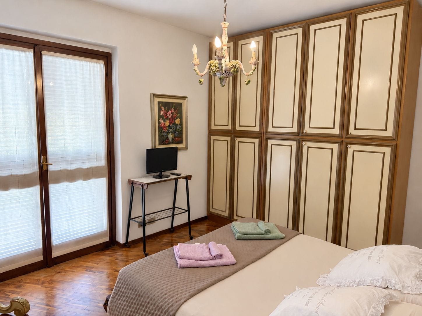 Une chambre à coucher avec un lit, un bureau avec un ordinateur, un lustre et une grande fenêtre avec des stores.