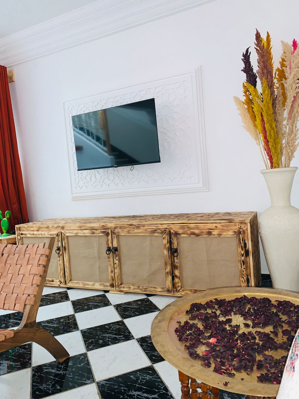 A room with a checkered floor, a television mounted on the wall, a wooden cabinet, and a vase with dried flowers.