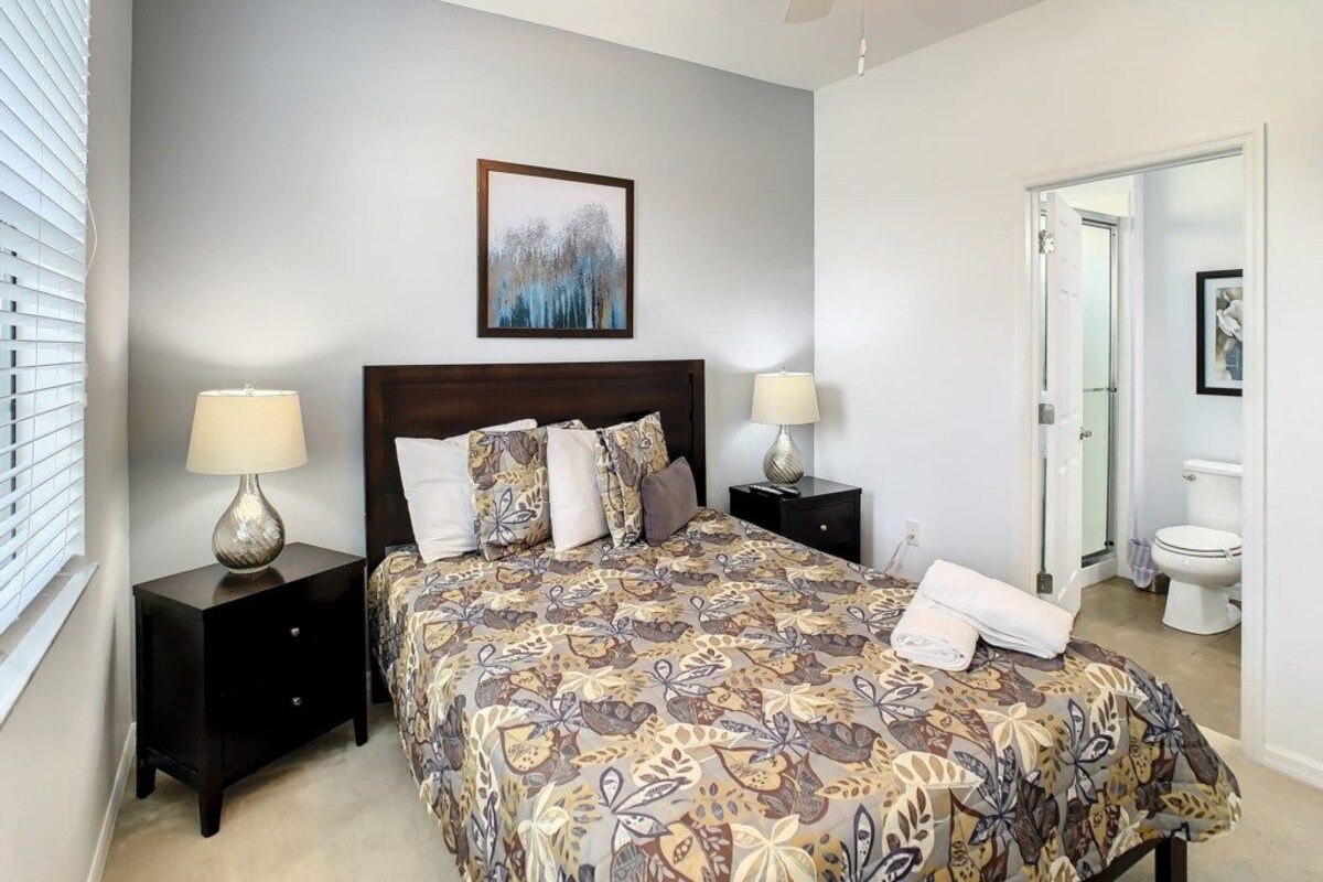 A bedroom with a bed, two bedside tables, a ceiling fan, a framed artwork above the bed, and a bathroom door in the background.