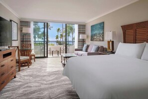 Habitación de hotel con una cama grande, un escritorio, una silla, un sofá y un balcón con vistas a palmeras.