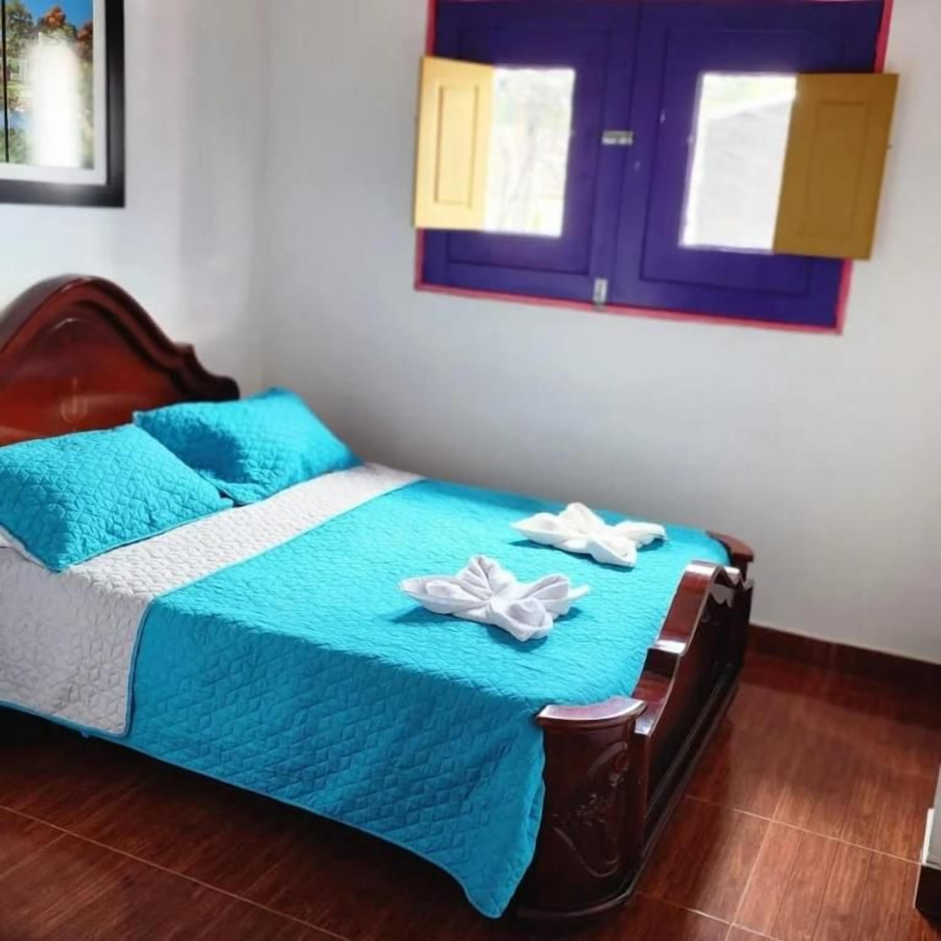 Una cama con colcha azul y almohadas blancas, cabecera de madera y ventana con marco morado y contraventanas amarillas.