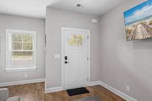 A hallway with a white door, a window with blinds, a black doormat, and a wall-mounted picture. - Your Brand New Beach Retreat! (Pawleys Island)