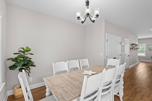 A dining area with a wooden table, white chairs, a chandelier, and a potted plant. - Your Brand New Beach Retreat! (Pawleys Island)