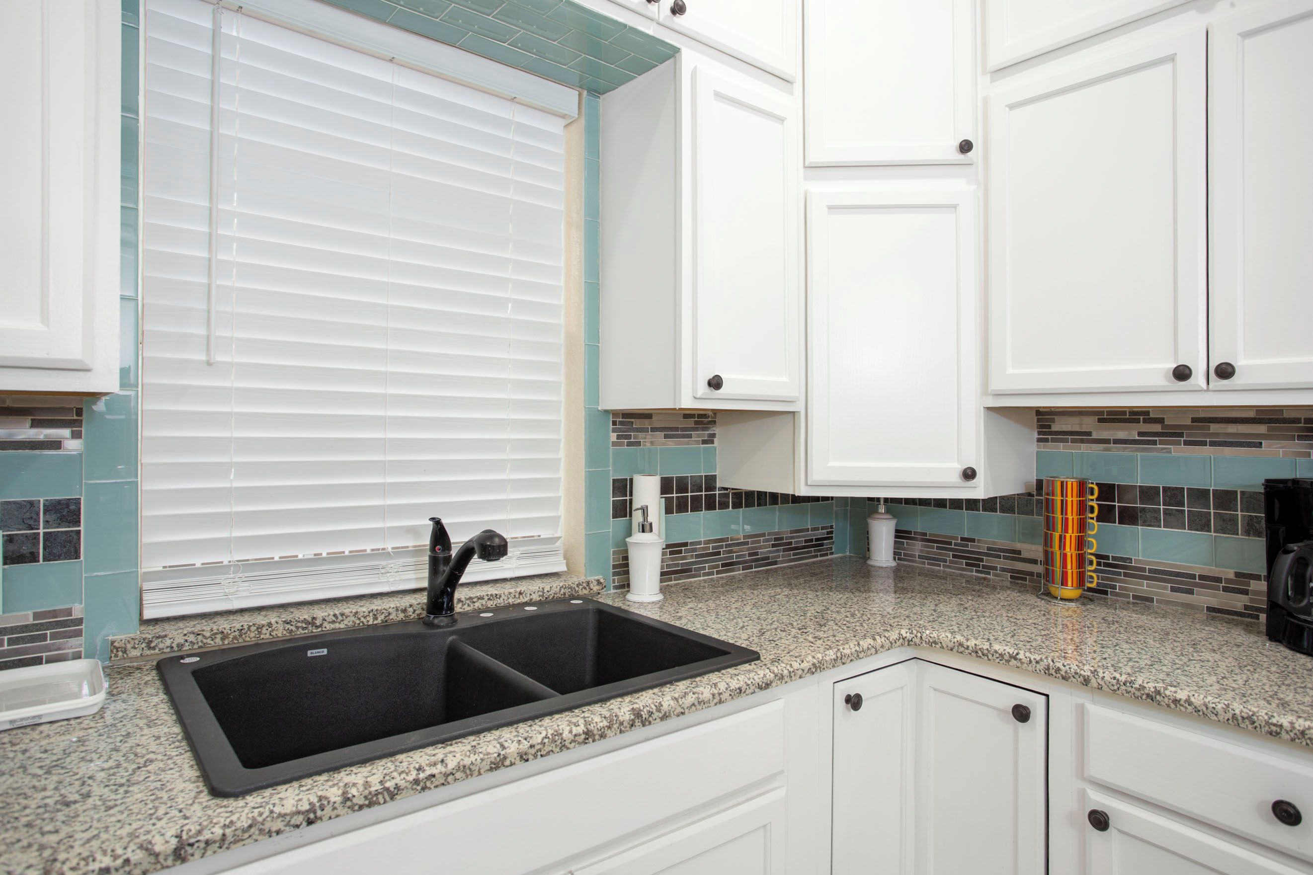 A kitchen with white cabinets, a black sink, and a window with blinds.