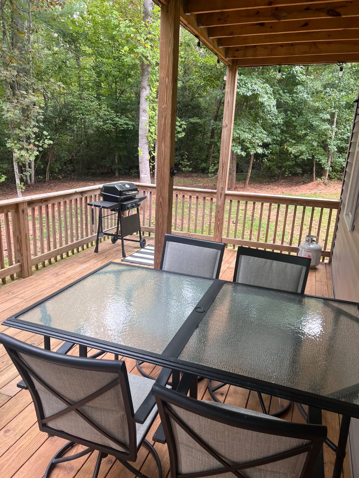A covered outdoor deck with a glass-top table and metal chairs, surrounded by a wooden railing.