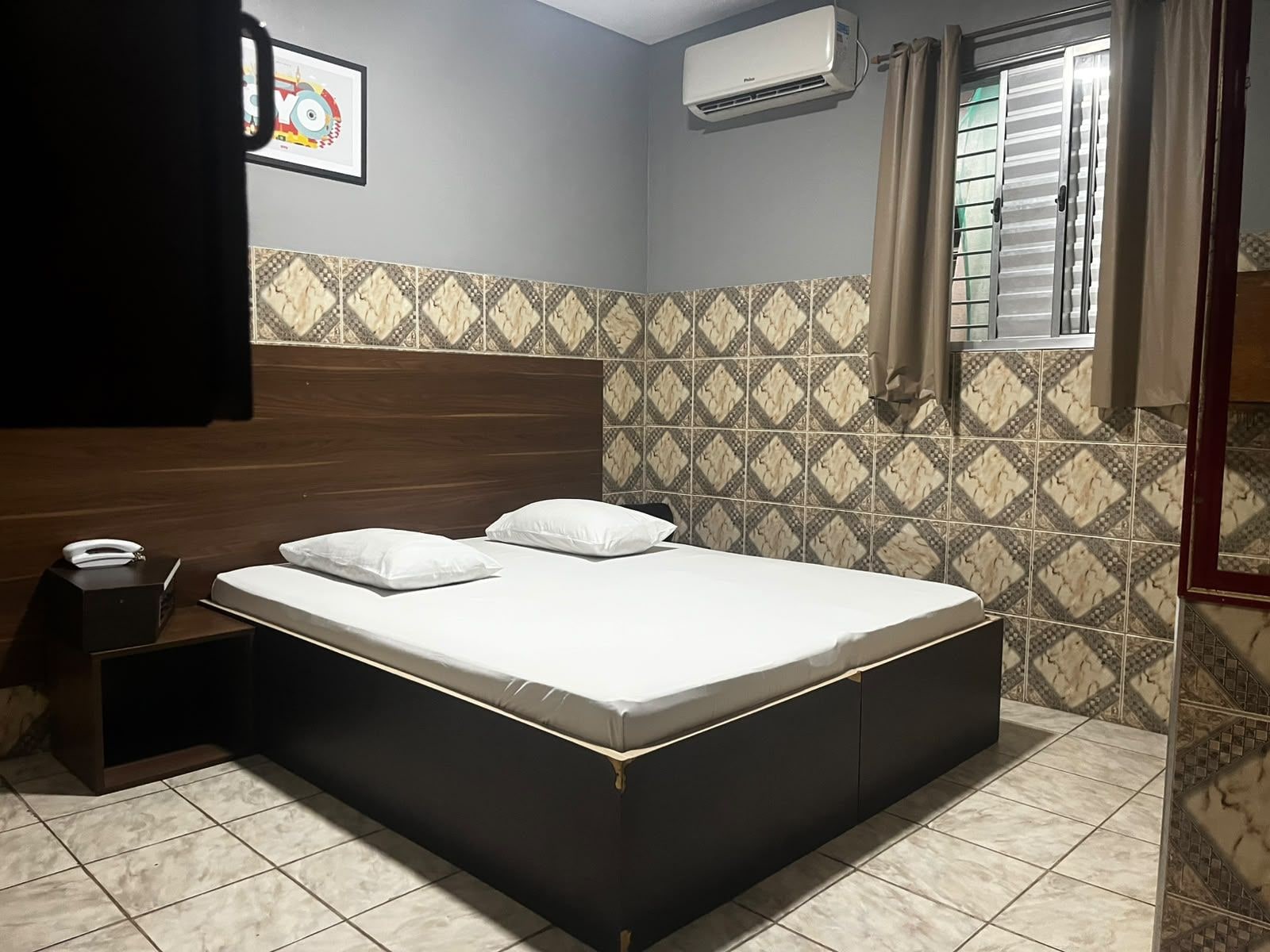 A bedroom with a bed, a nightstand, an air conditioning unit, and patterned tiles.