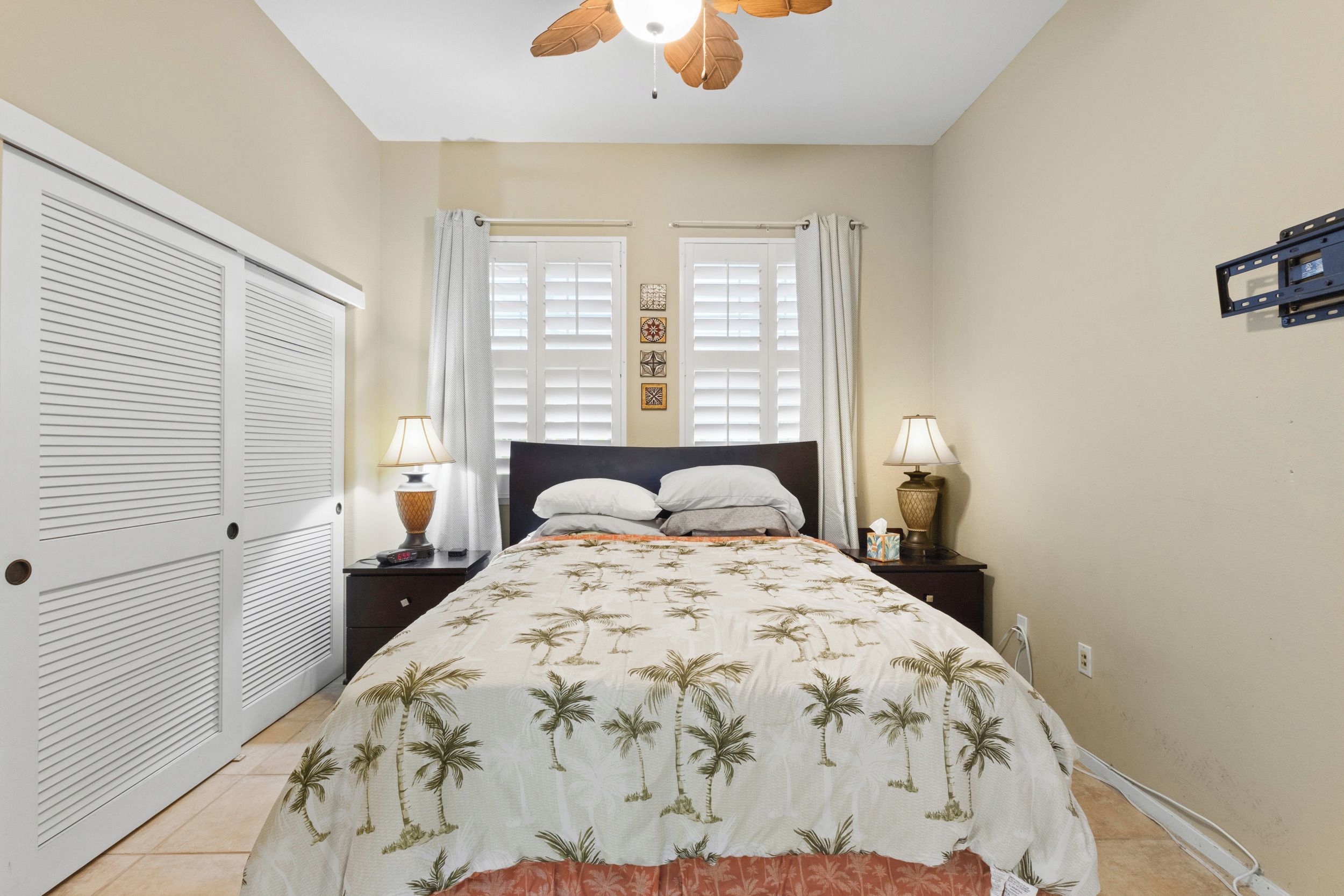 Une chambre à coucher comprenant un lit, deux tables de chevet, un ventilateur de plafond et une fenêtre avec des stores.