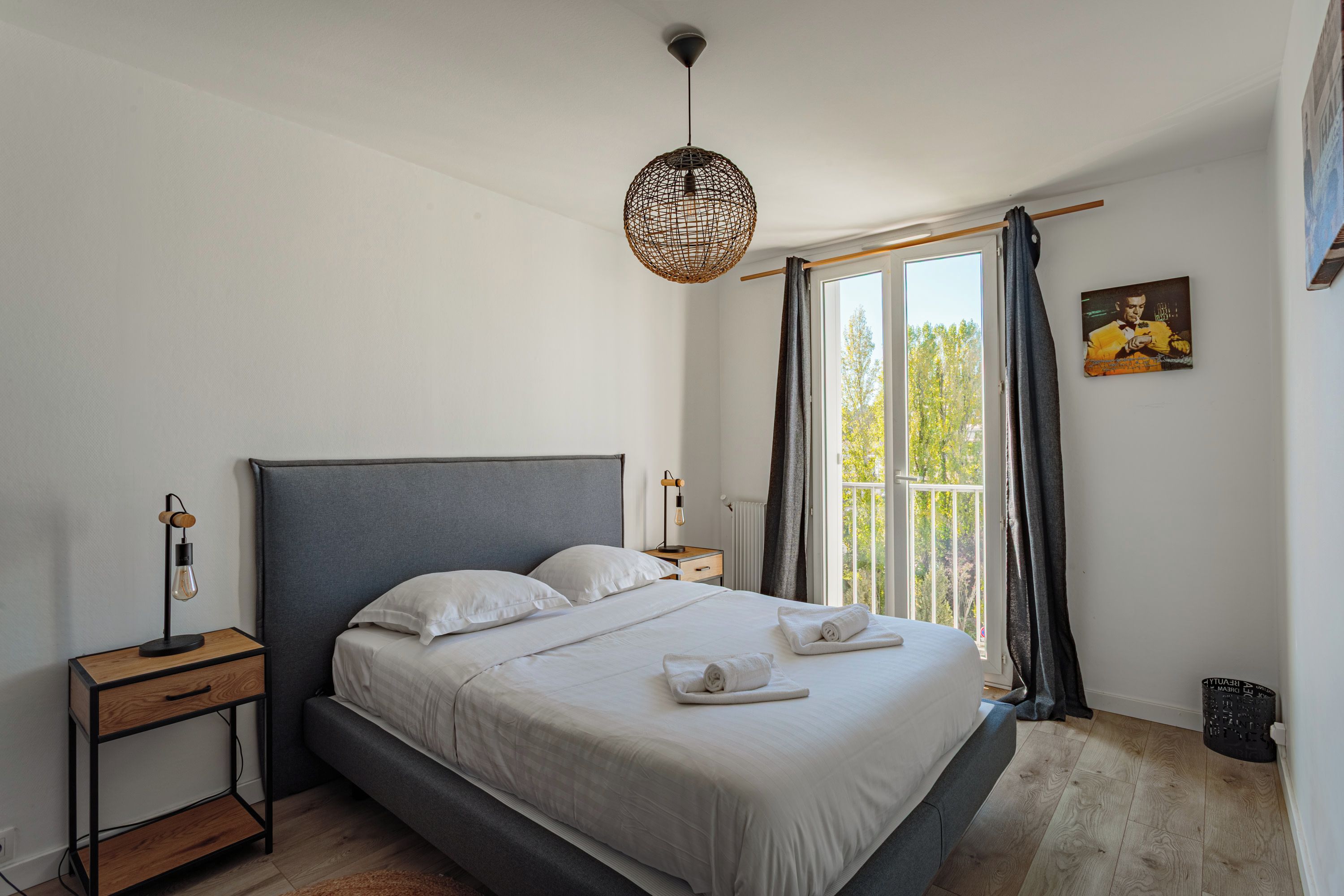 A bedroom with a bed, bedside tables, a hanging light fixture, a window with curtains, and a wall-mounted picture.