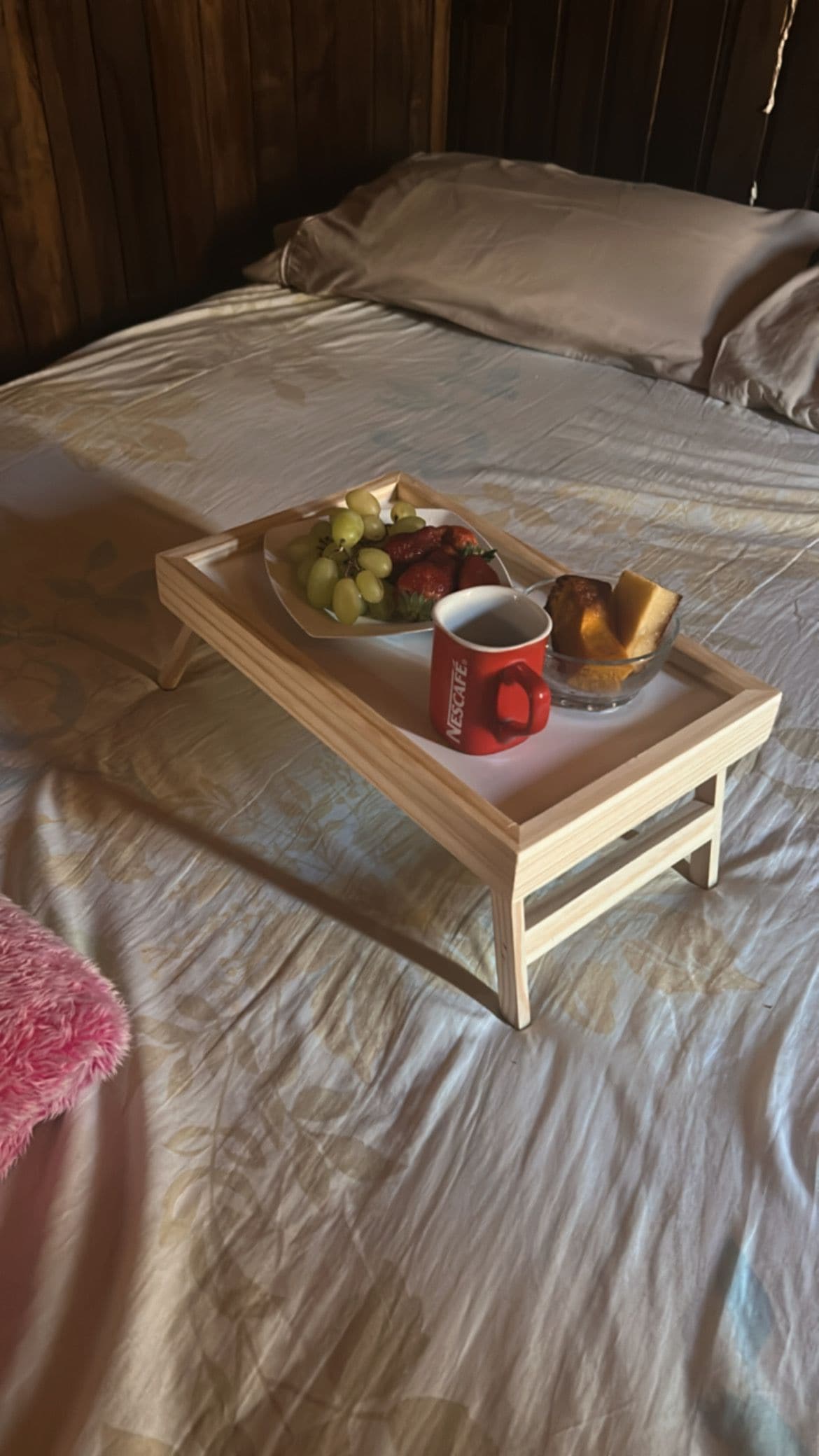Een bed met een houten dienblad waarop een bord met fruit, een rode mok en een glazen schaal staan.