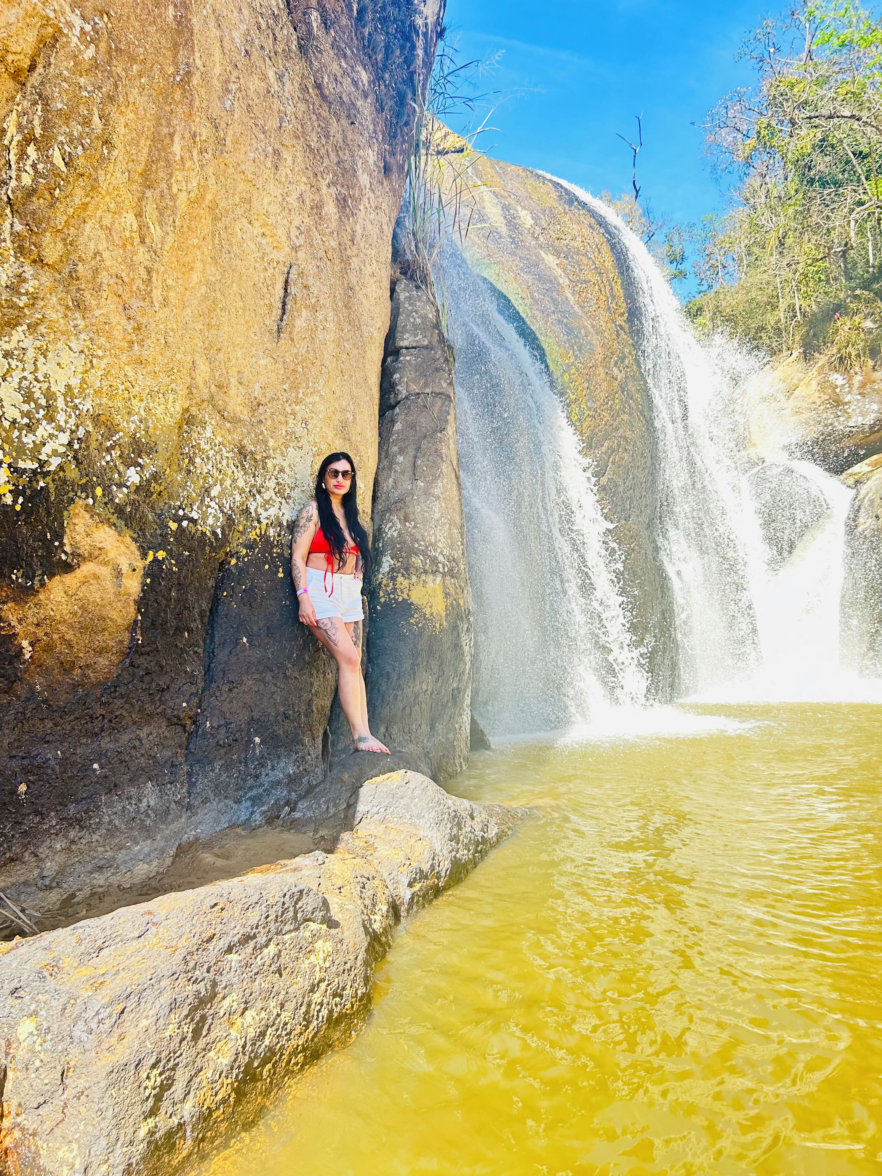 Uma pessoa parada perto de uma cachoeira, com terreno rochoso e um lago de água.