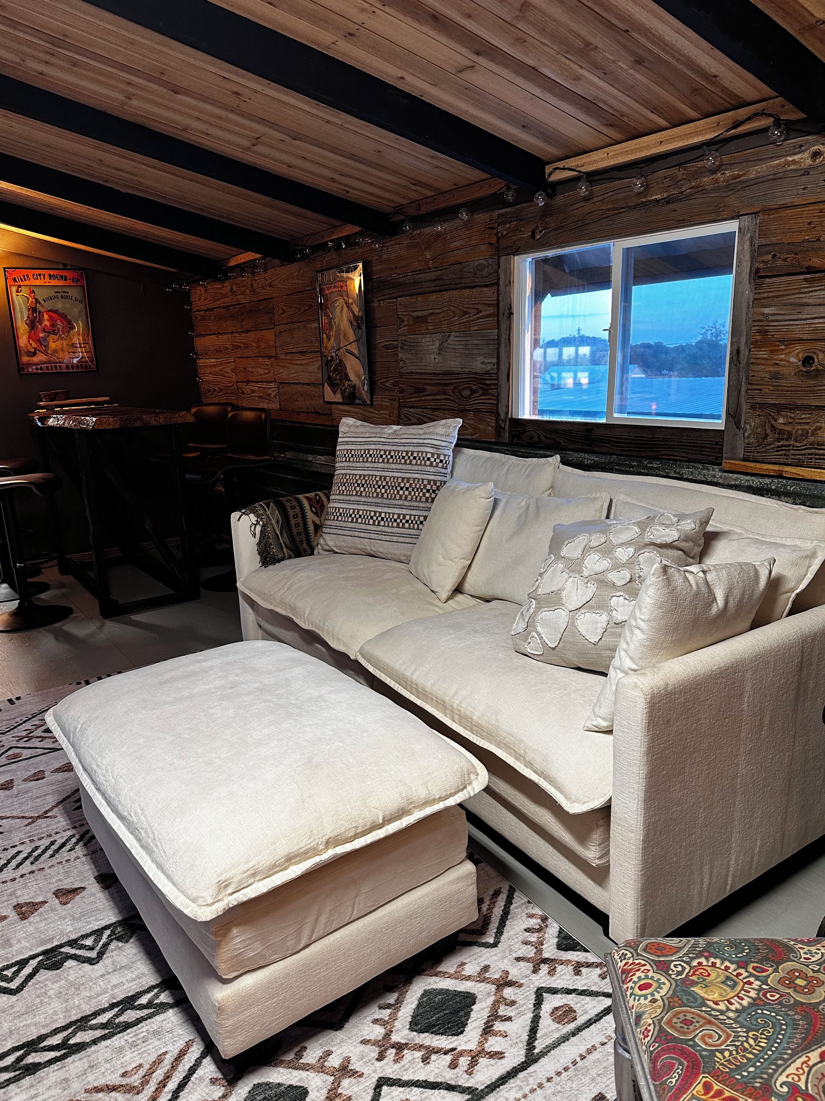 A cozy room with a wooden ceiling, a large window, a sofa with patterned cushions, and a patterned rug.