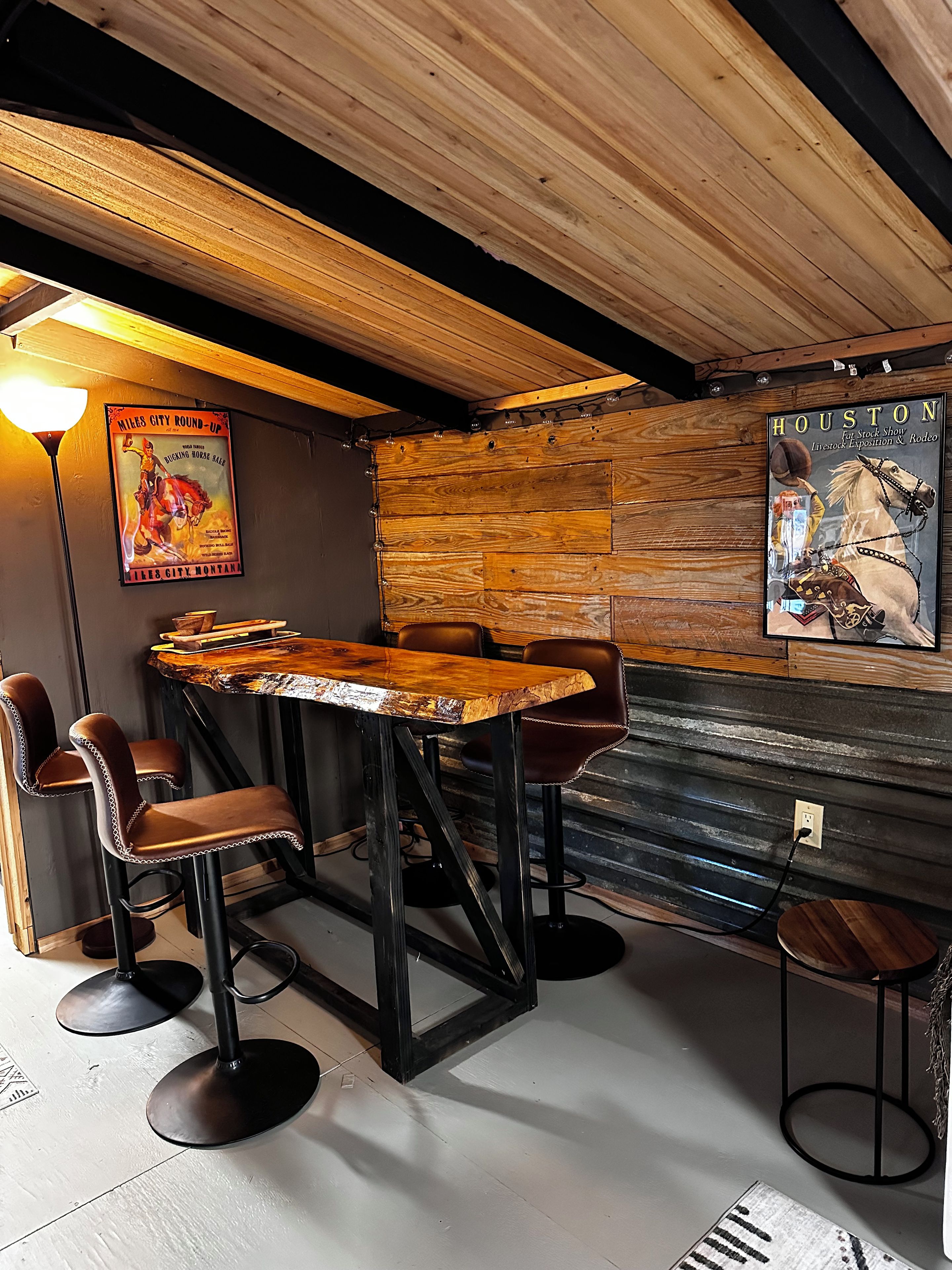 A rustic bar area with wooden tables and stools, a poster of a rodeo event, and a wall-mounted sign.