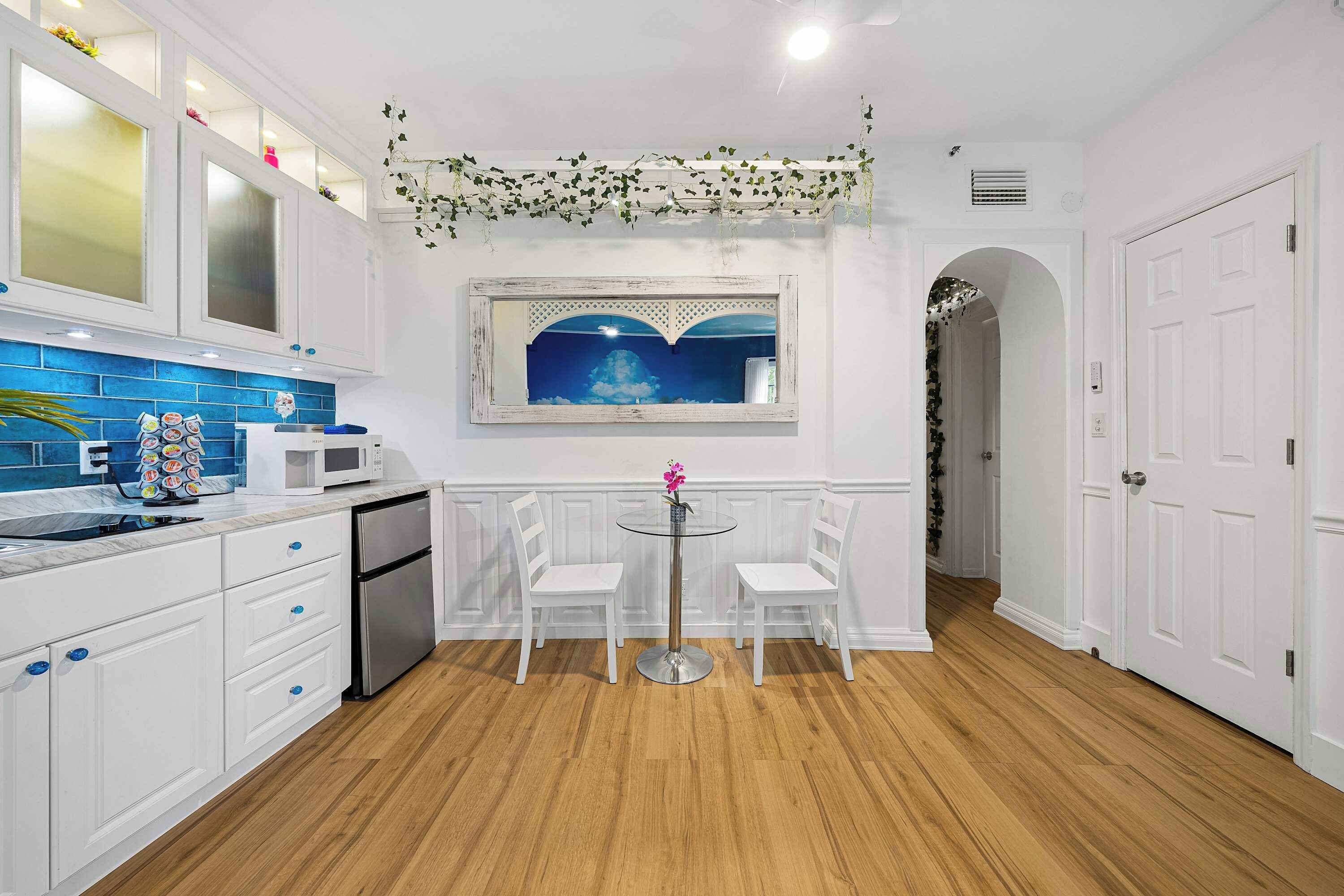 Una cocina moderna con armarios blancos, un salpicadero de azulejos azules y una zona de comedor con dos sillas blancas y una mesa de vidrio.