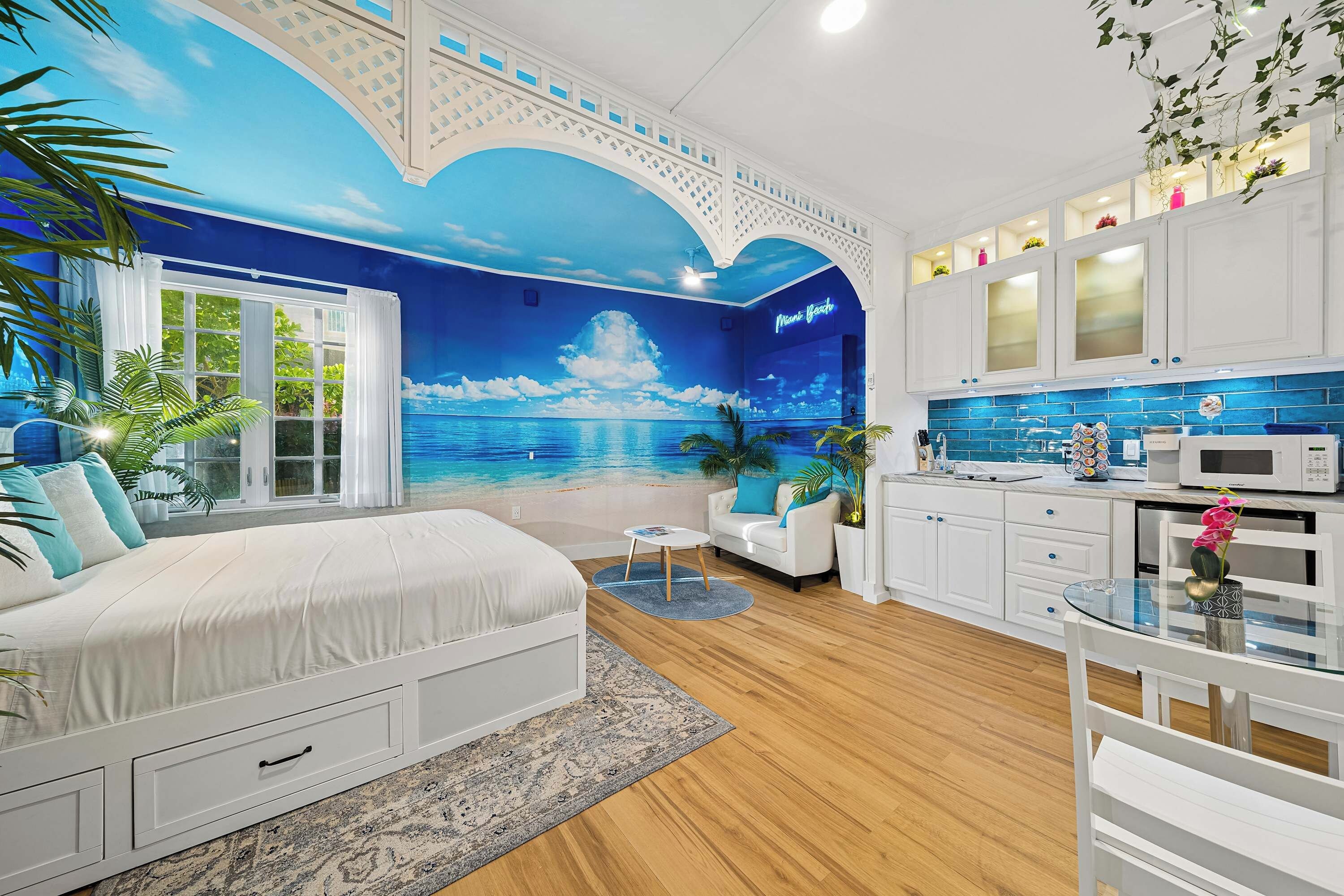 Un dormitorio con una cama grande, una sala de estar con sofá y una mesita, y una cocina con armarios blancos y salpicadero azul.