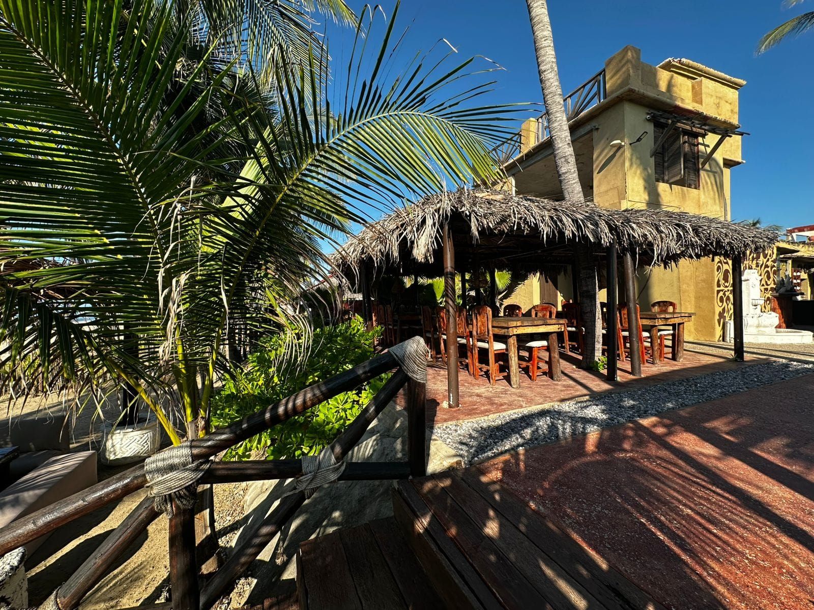 A thatched-roof outdoor seating area with wooden furniture, surrounded by palm trees and a building with a yellow facade.