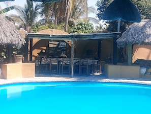 Un espace aménagé au bord de la piscine, comprenant un coin salon couvert, un barbecue et un parasol en chaume.