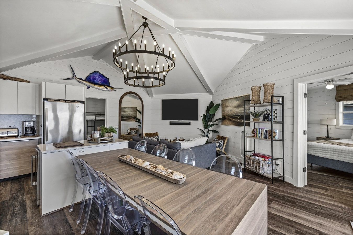 A modern kitchen with a central island, a dining area with clear chairs, and a living space with a sofa and a large window.