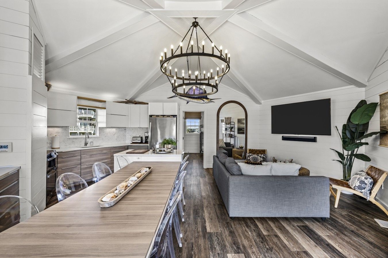 A modern kitchen with a central island, a dining area with a long table, and a living room with a sofa and a TV.