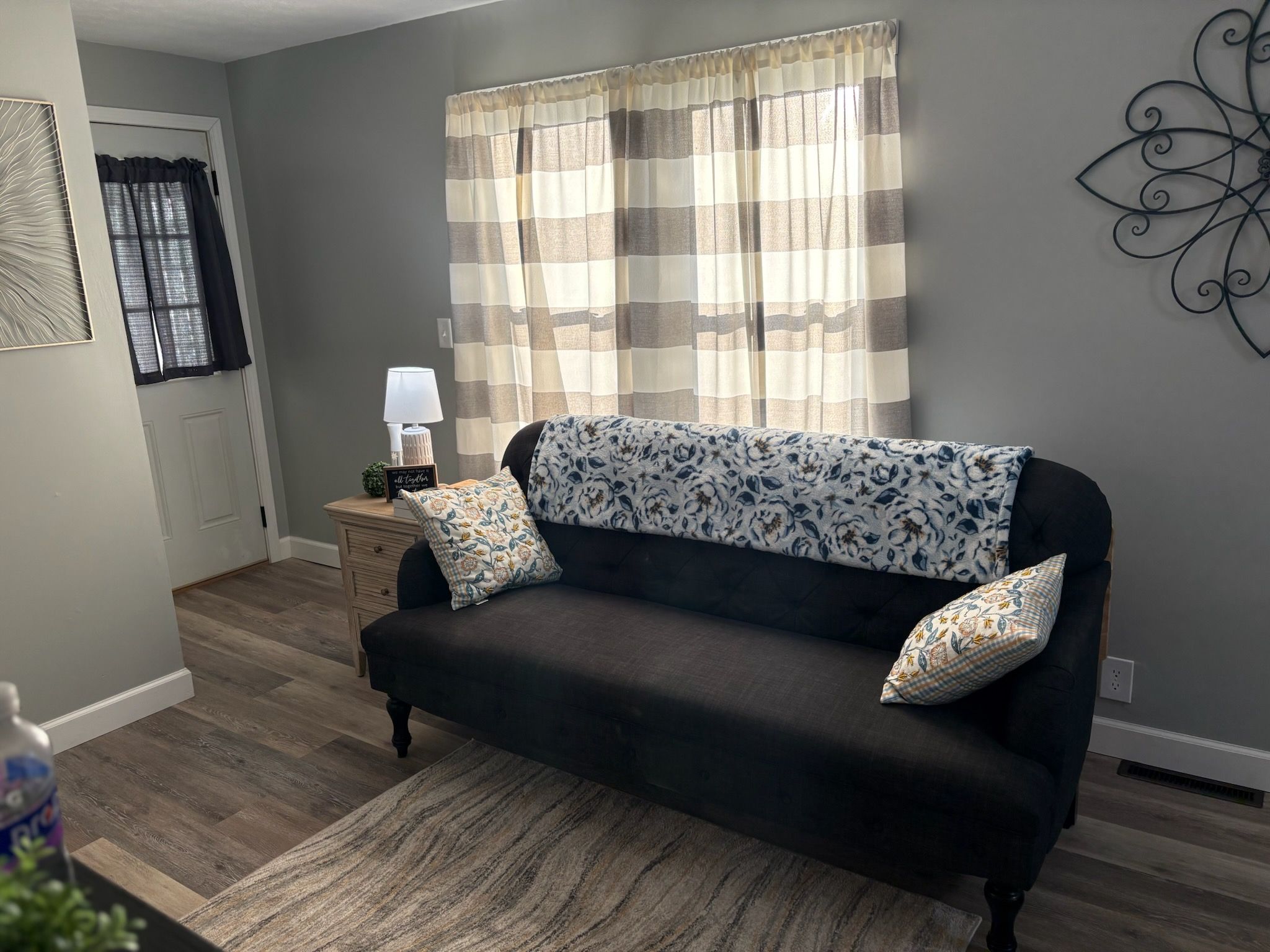 A living room with a dark sofa, patterned cushions, a lamp, and a window with curtains.