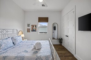 Een slaapkamer met een bed, nachtkastje, lamp, raam, spiegel en een aan de muur bevestigde televisie.