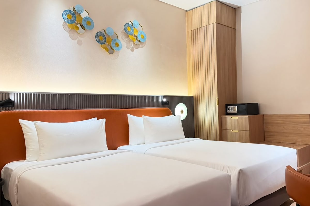 A hotel room with two beds, a wooden wardrobe, a desk with a chair, and a wall-mounted light fixture.