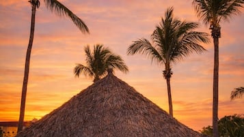 Une structure à toit de chaume dotée d’un espace extérieur pour s’asseoir, entourée de palmiers, au coucher du soleil.
