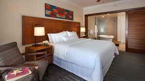 Habitación de hotel con una cama grande, cabecera de madera, dos mesitas de noche con lámparas, una silla y vistas al baño.