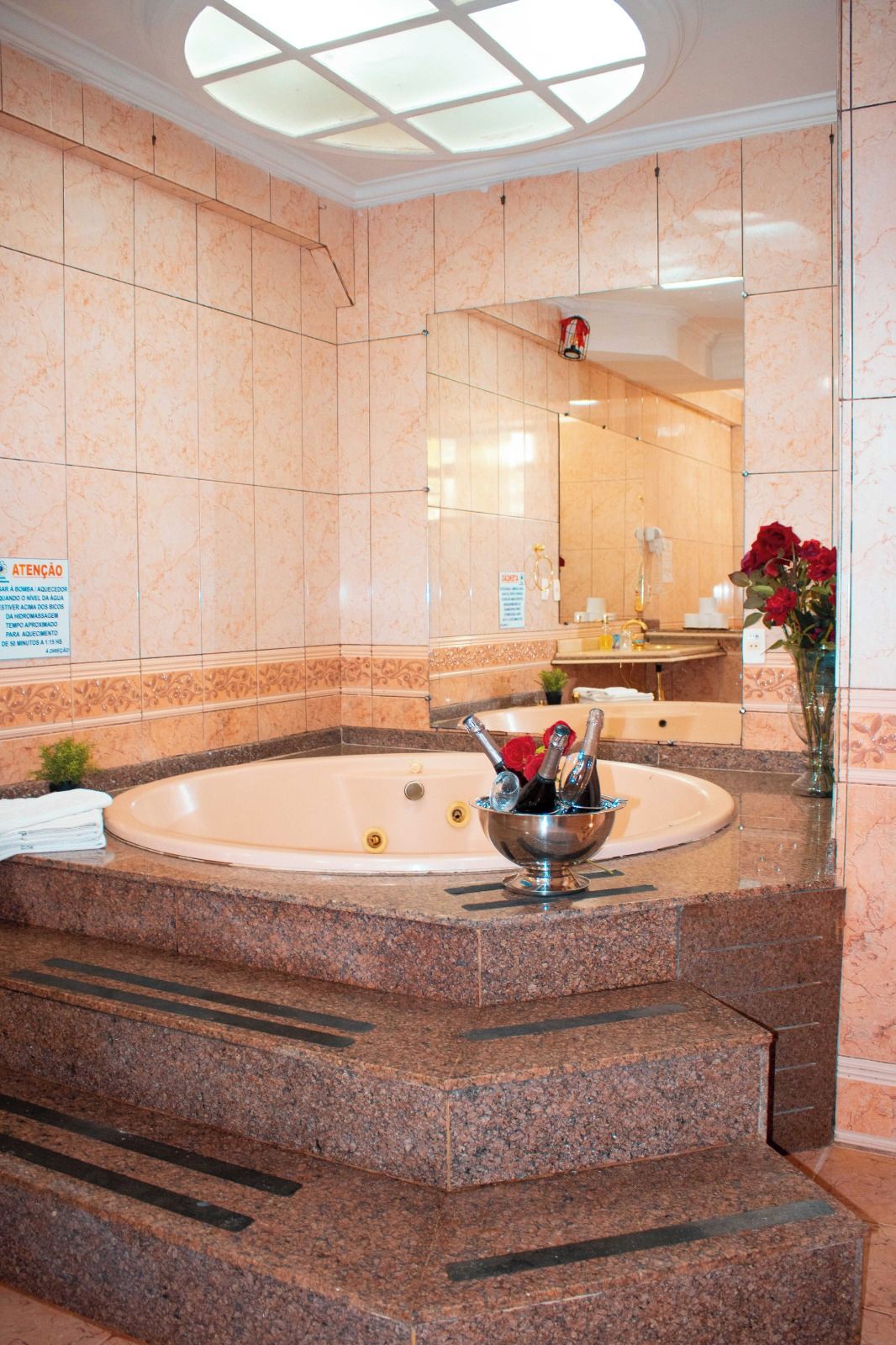 Casa de banho de hotel com parede revestida a mármore, um espelho grande, uma banheira e uma tigela de metal com garrafas e uma flor.