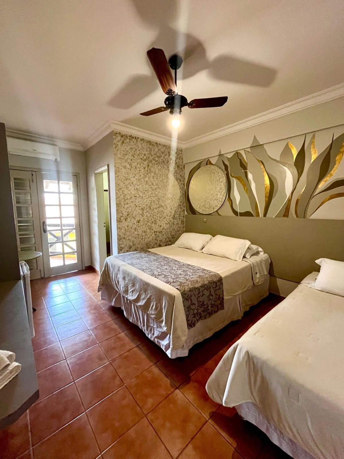 Quarto de hotel com cama, mesa de cabeceira, cadeira, ventilador de teto e painel decorativo na parede.