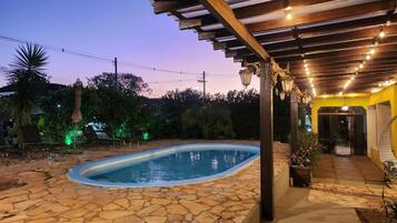 A swimming pool with stone flooring, surrounded by a covered patio area with hanging lights.