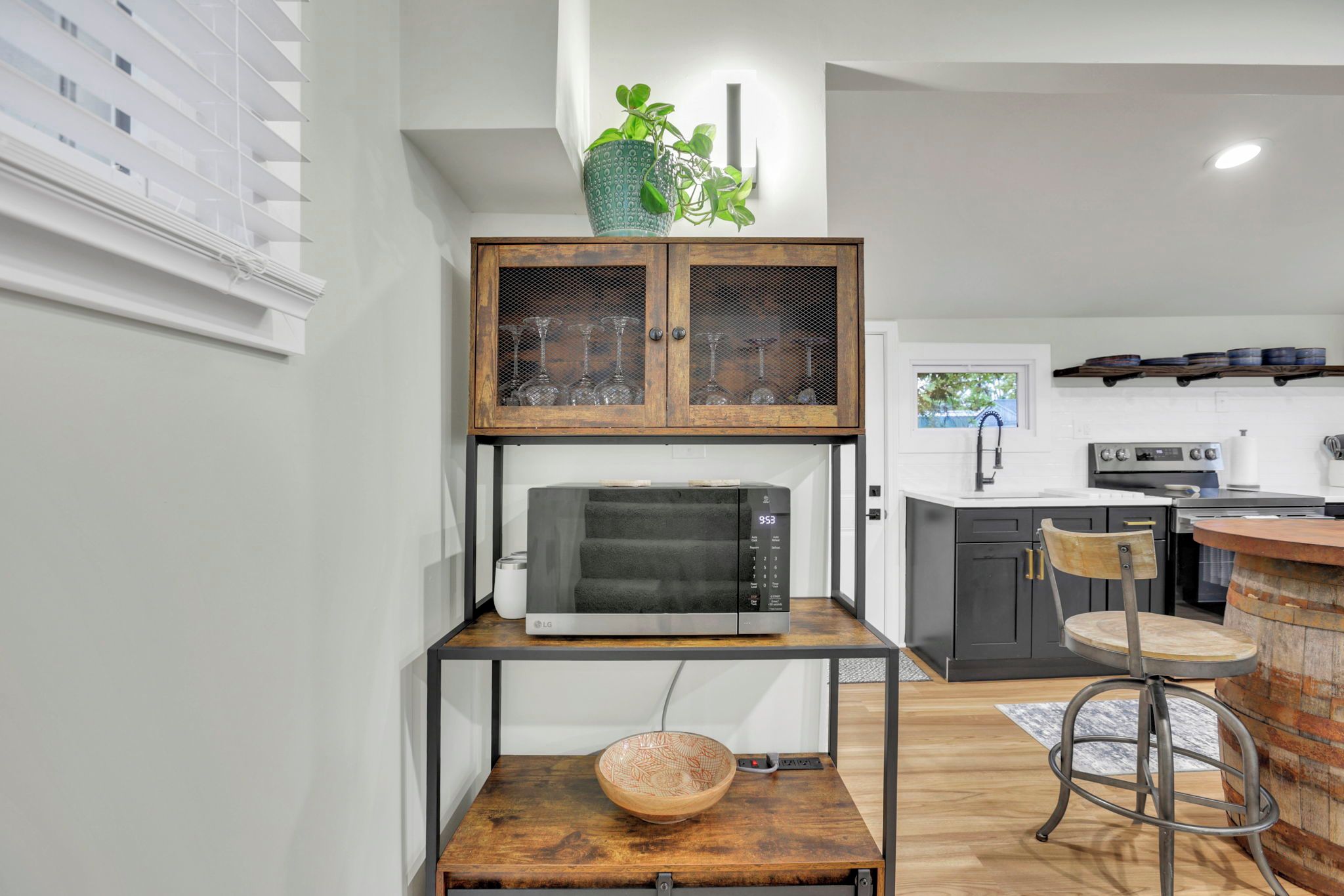 A compact kitchen with a microwave, a wooden cabinet with glass doors, a bar stool, and a potted plant.
