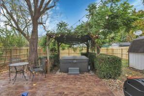 A backyard with a hot tub, a pergola with string lights, a wooden deck, and a barbecue grill.
