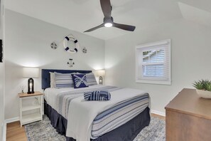 A bedroom with a bed, bedside lamps, a ceiling fan, a window with blinds, and nautical-themed wall decor.