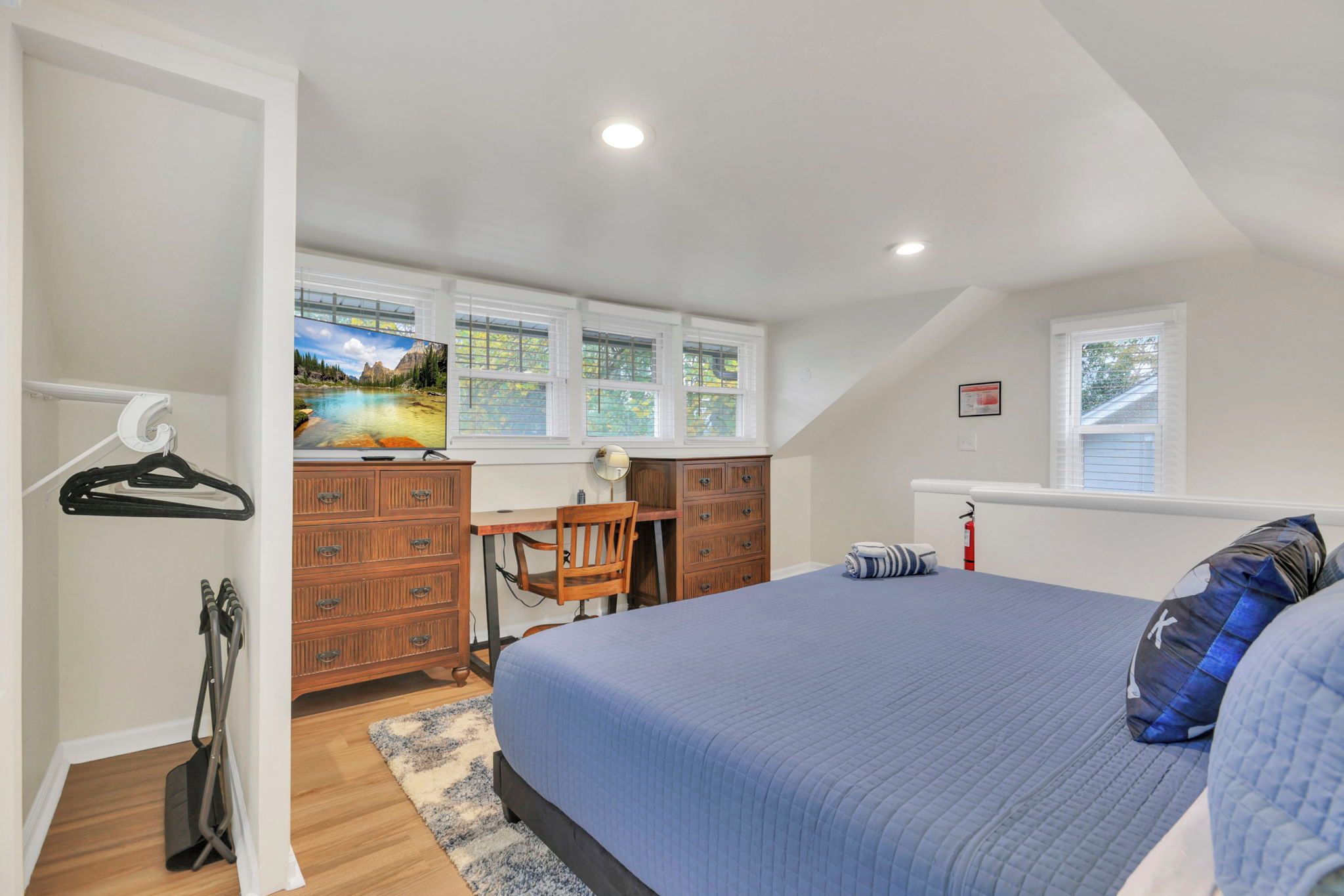 A bedroom with a bed, a desk, and a chair. There is a window with blinds, a fire extinguisher, and a coat rack.