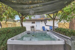 A hot tub with a wooden deck and a covered area, surrounded by greenery and a house in the background.
