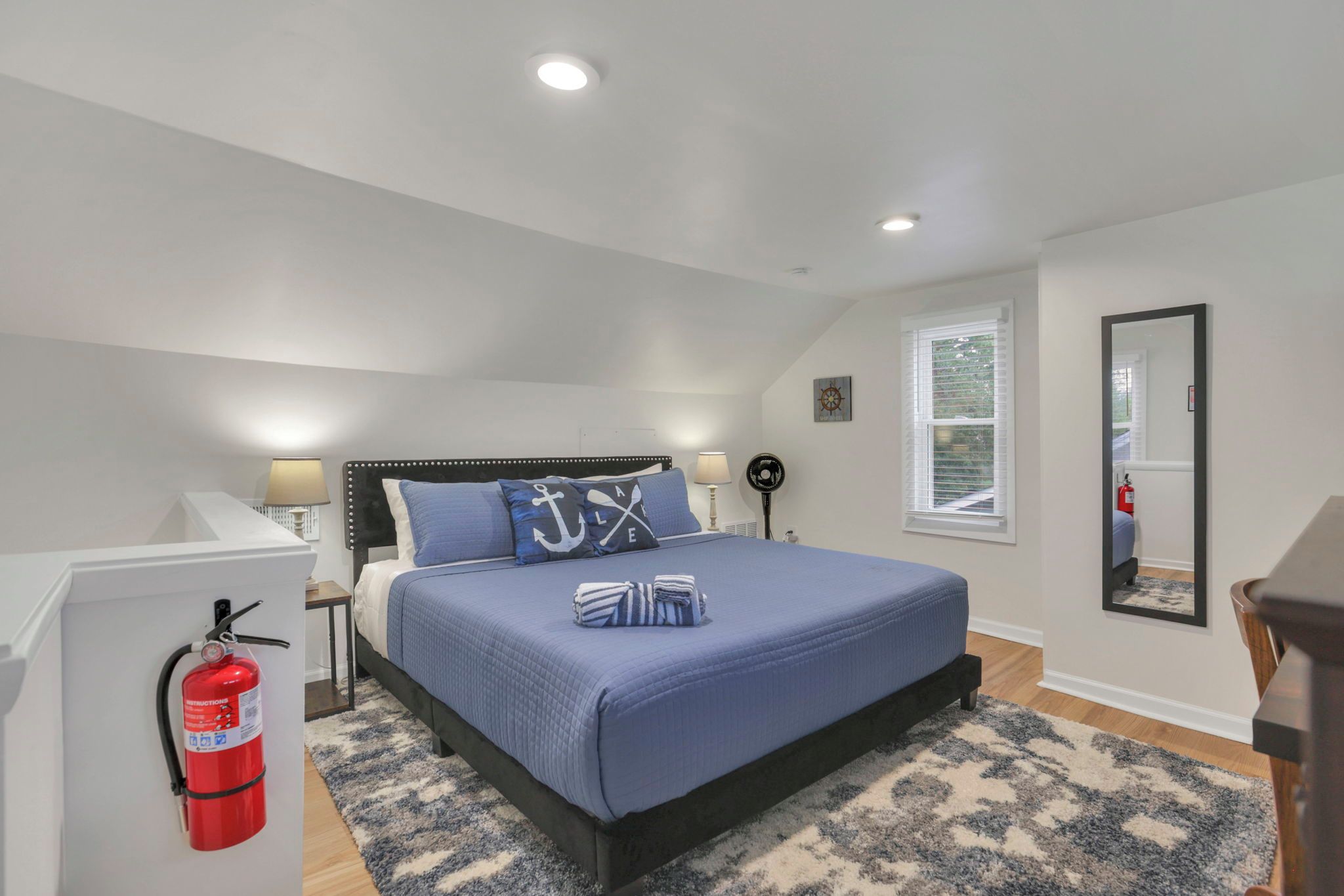 A bedroom with a bed, bedside lamps, a fire extinguisher, a window with blinds, and a mirror on the wall.