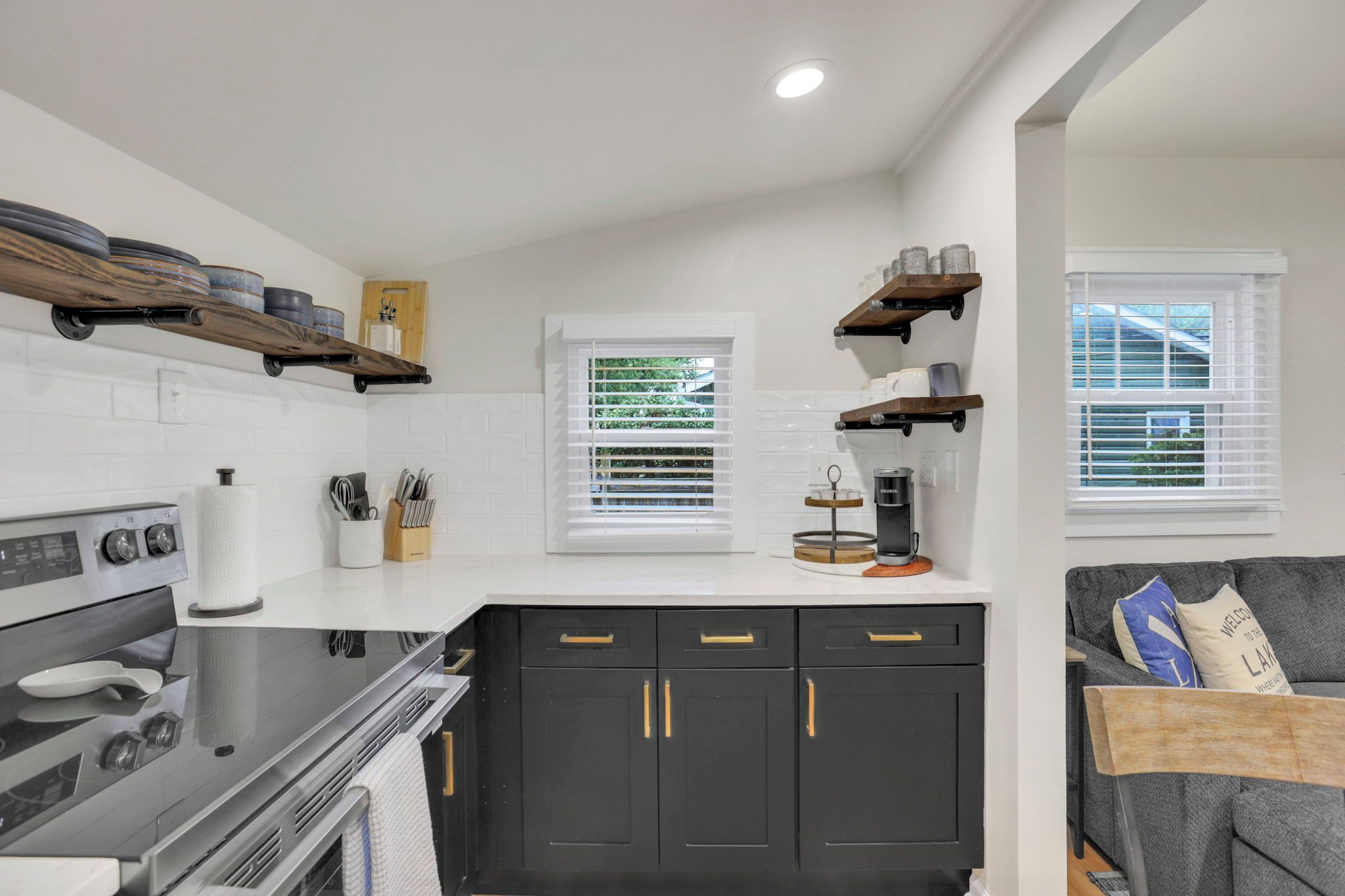 A compact kitchen with a stove, oven, and black cabinets with gold handles. There are two windows with blinds, a coffee maker on the counter, and a gray couch in the living area.