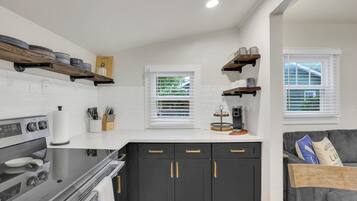 A compact kitchen with a stove, oven, and black cabinets with gold handles. There are two windows with blinds, a coffee maker on the counter, and a gray couch in the living area.
