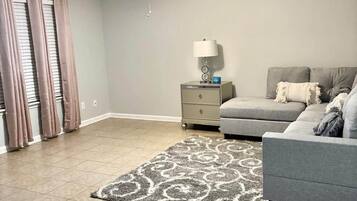 A bedroom with a ceiling fan, a nightstand with a lamp, a grey sofa, and a patterned rug.