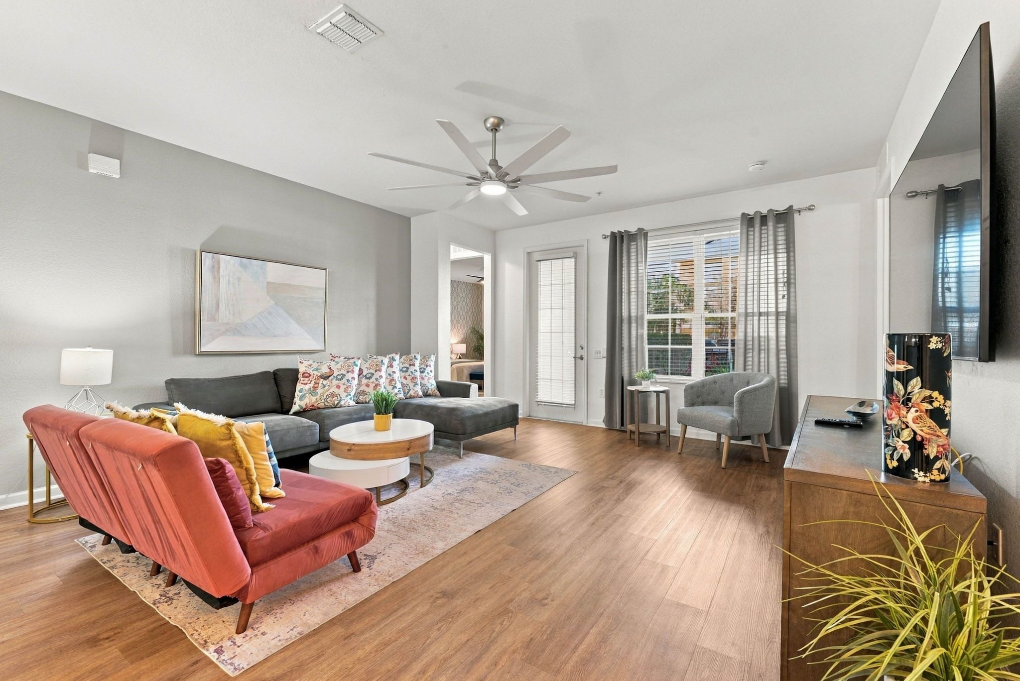 Un salon moderne avec un canapé gris, un fauteuil rouge, une table basse et un autre fauteuil gris. On y trouve un ventilateur de plafond, une fenêtre avec des rideaux et un téléviseur fixé au mur.