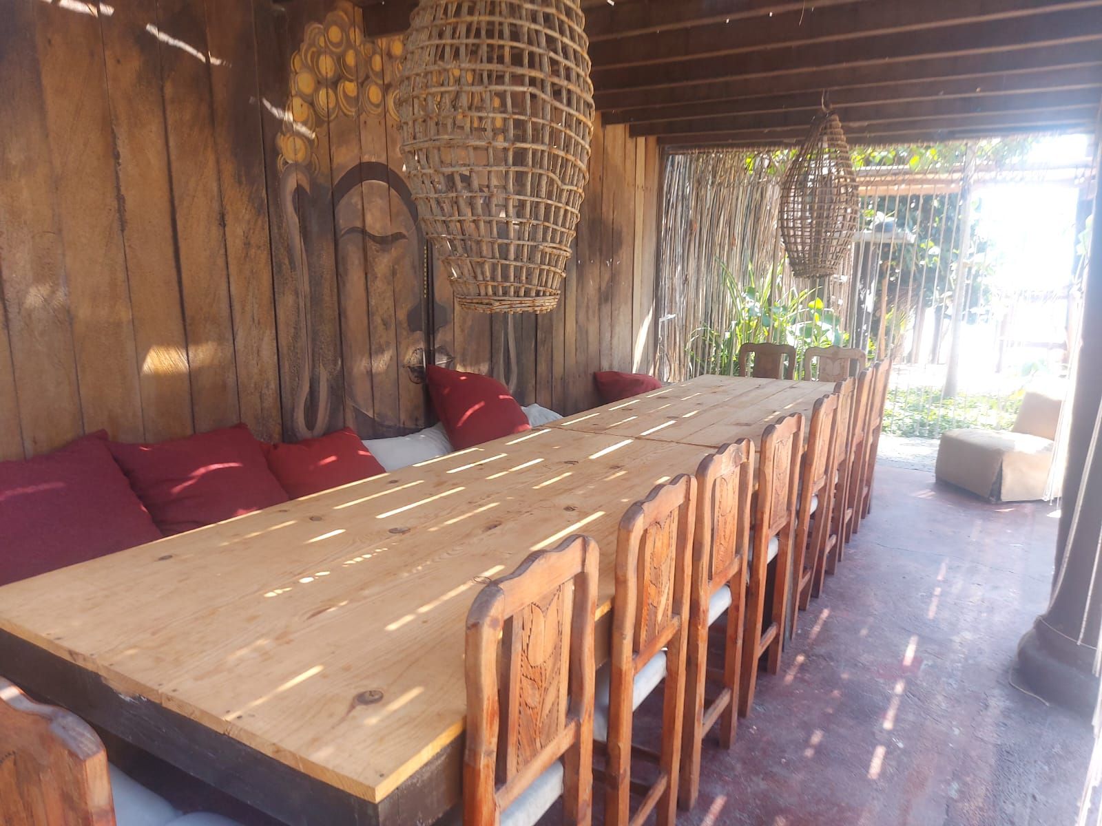 A wooden dining table with chairs, a woven ceiling, and a view of greenery outside.