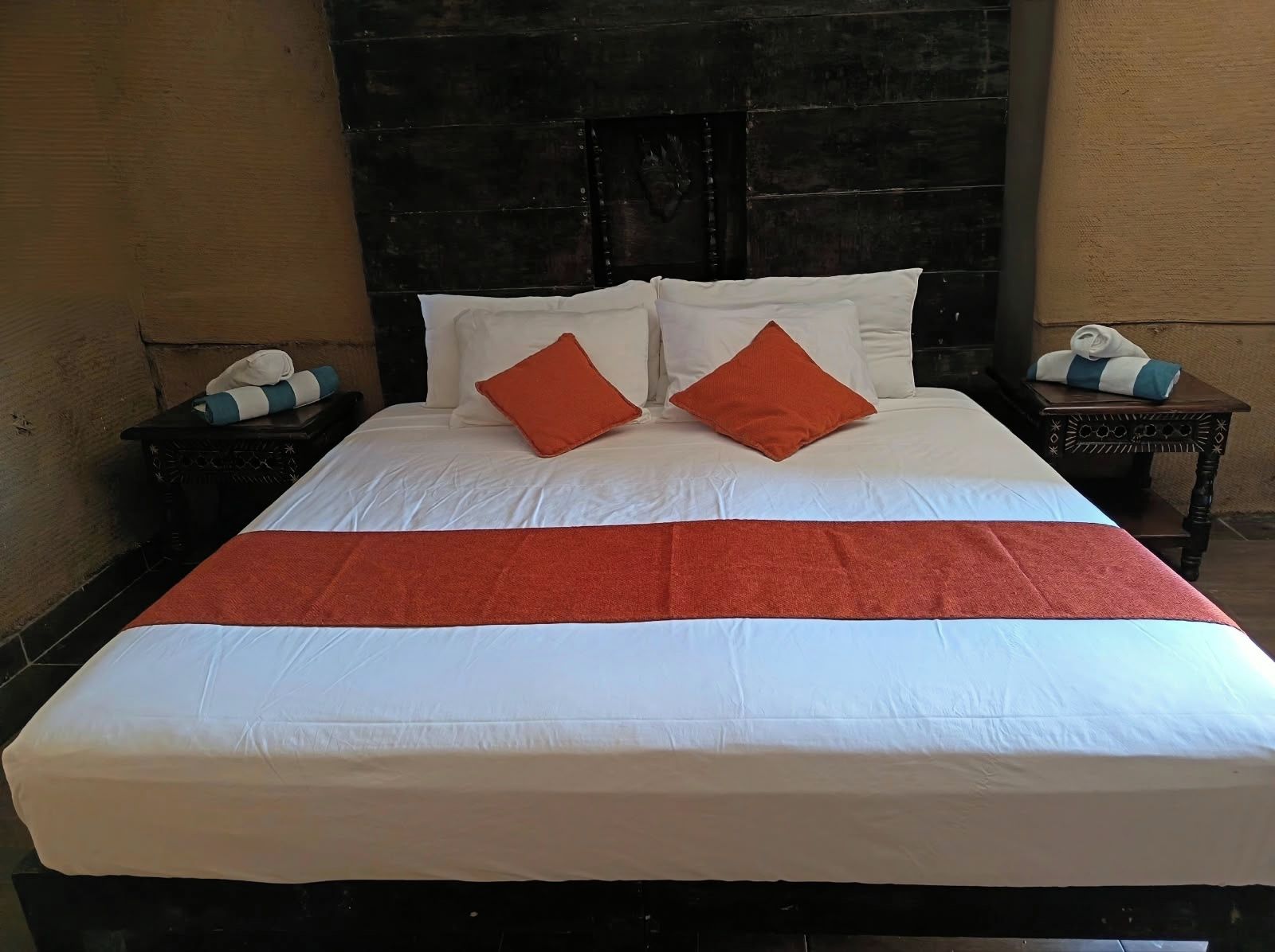 Een bed met witte beddengoed en twee oranje kussens, een houten hoofdbord en een nachtkastje met een handdoek en een klein tafeltje.