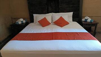 Een bed met witte beddengoed en twee oranje kussens, een houten hoofdbord en een nachtkastje met een handdoek en een klein tafeltje.