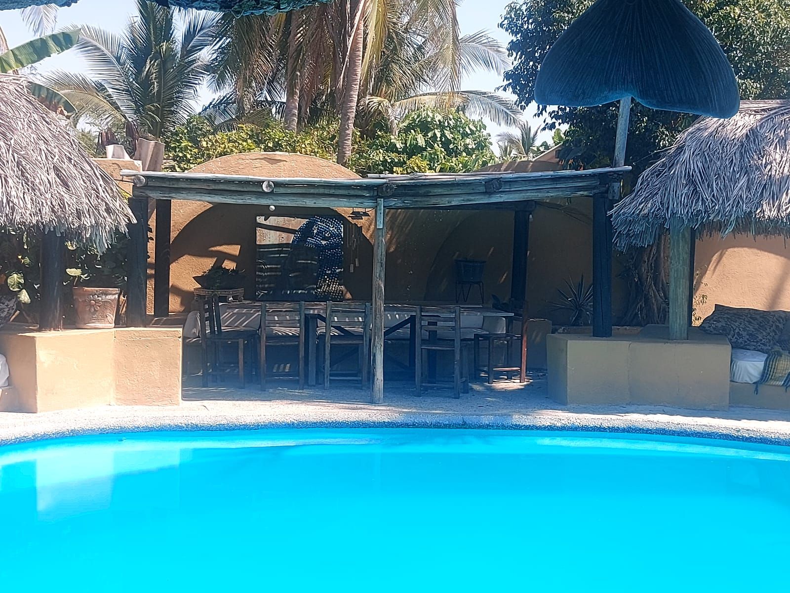 Un espace aménagé au bord de la piscine, comprenant un coin salon couvert, un barbecue et un parasol en chaume.