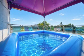 Una piscina con superficie de agua cristalina y azul, rodeada por una barandilla de vidrio, bajo un toldo morado.