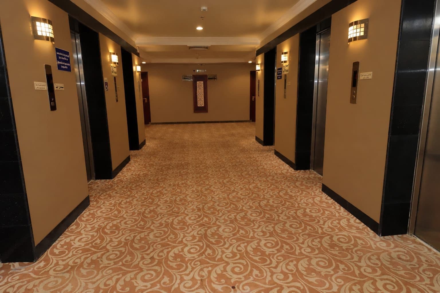 Un corridoio d'albergo con pavimenti rivestiti di moquette, porte dell'ascensore e luci a parete.